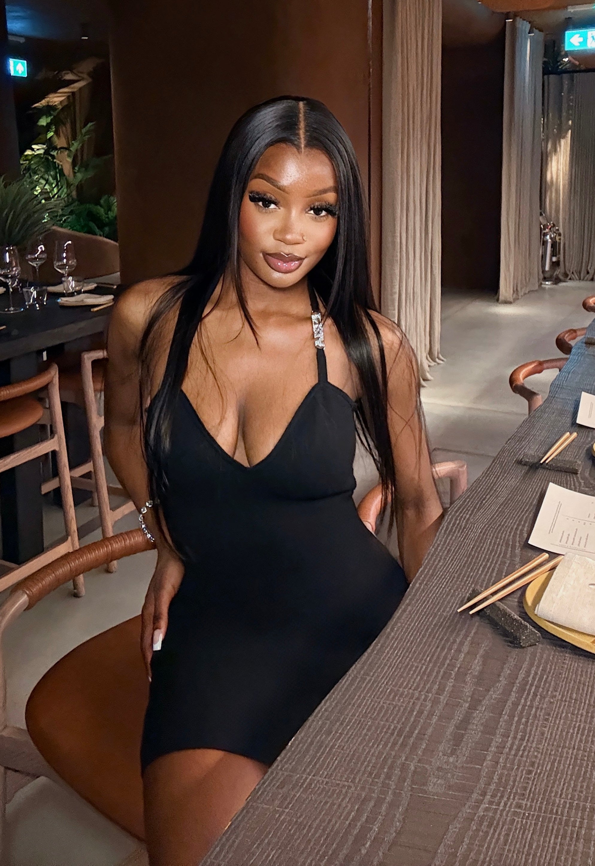 A woman with long straight black hair poses in a black halter-neck mini dress while seated at a table in a restaurant or lounge setting.