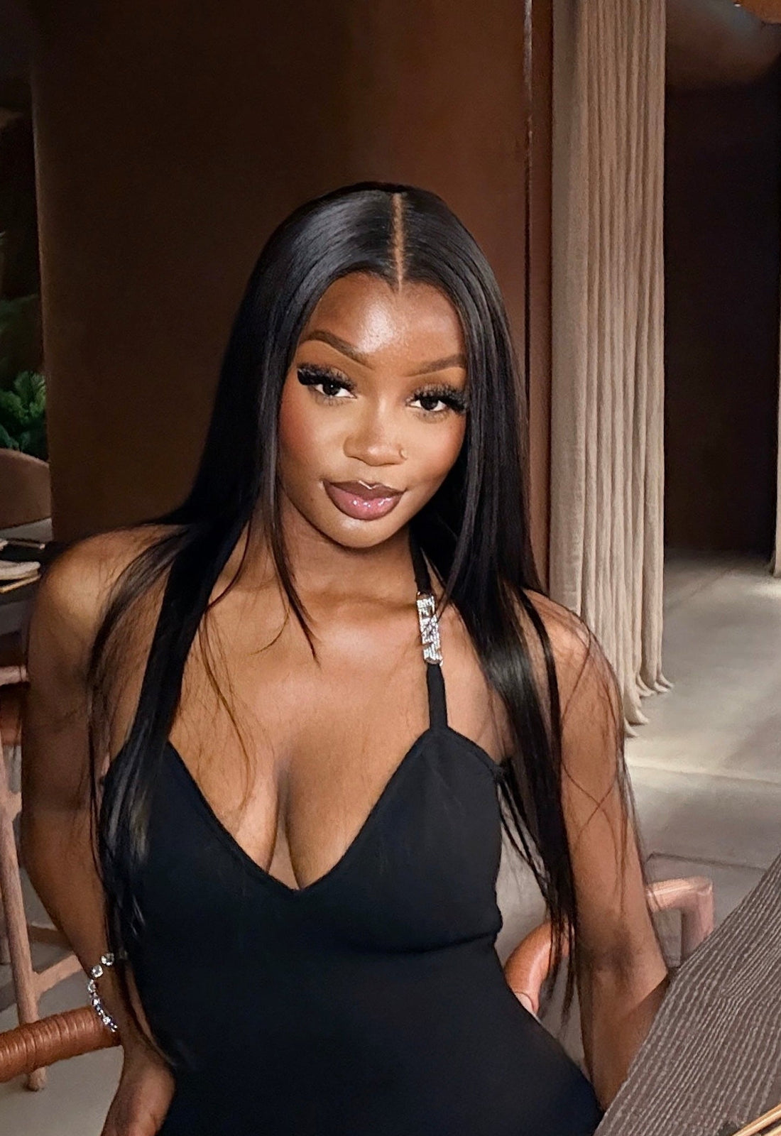 A woman with long straight black hair poses in a black halter-neck mini dress while seated at a table in a restaurant or lounge setting.