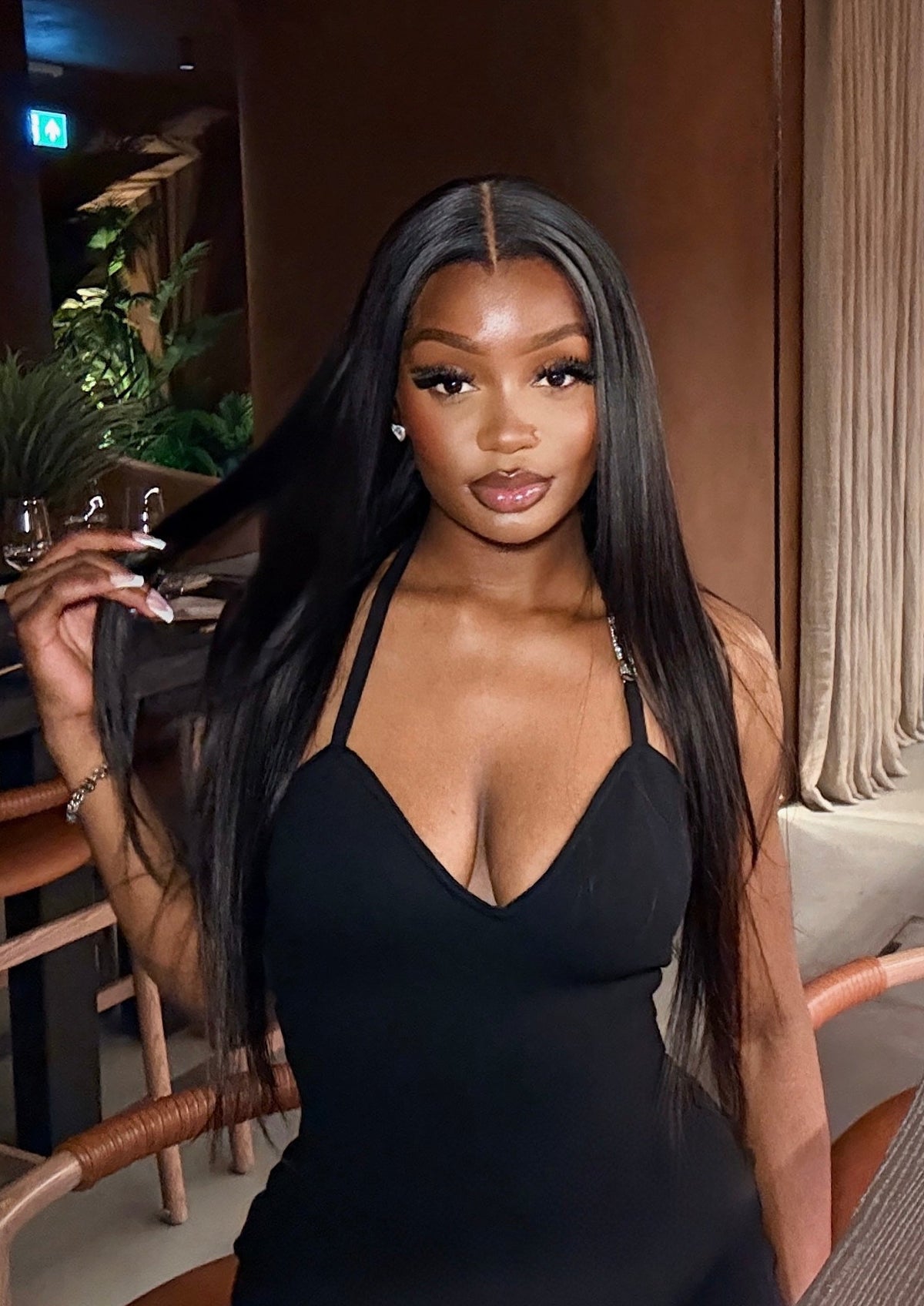 A woman with long, straight black hair wearing a black mini dress with a deep V-neckline, posing in a modern indoor setting with warm lighting.