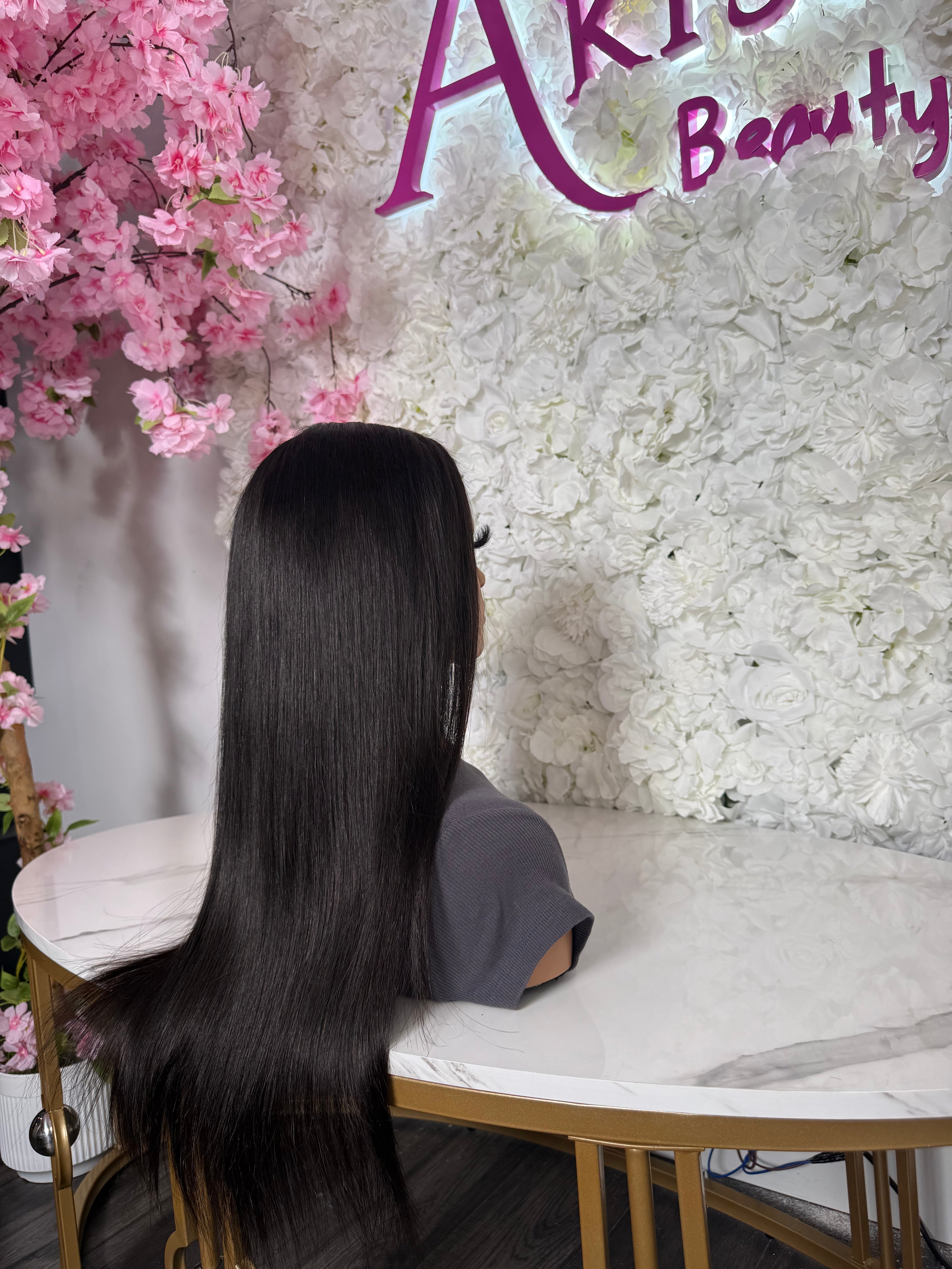 A long, straight, glossy black wig displayed on a mannequin head, shown from the back in a salon-like setting with a white floral wall, pink cherry blossoms, and a marble table.