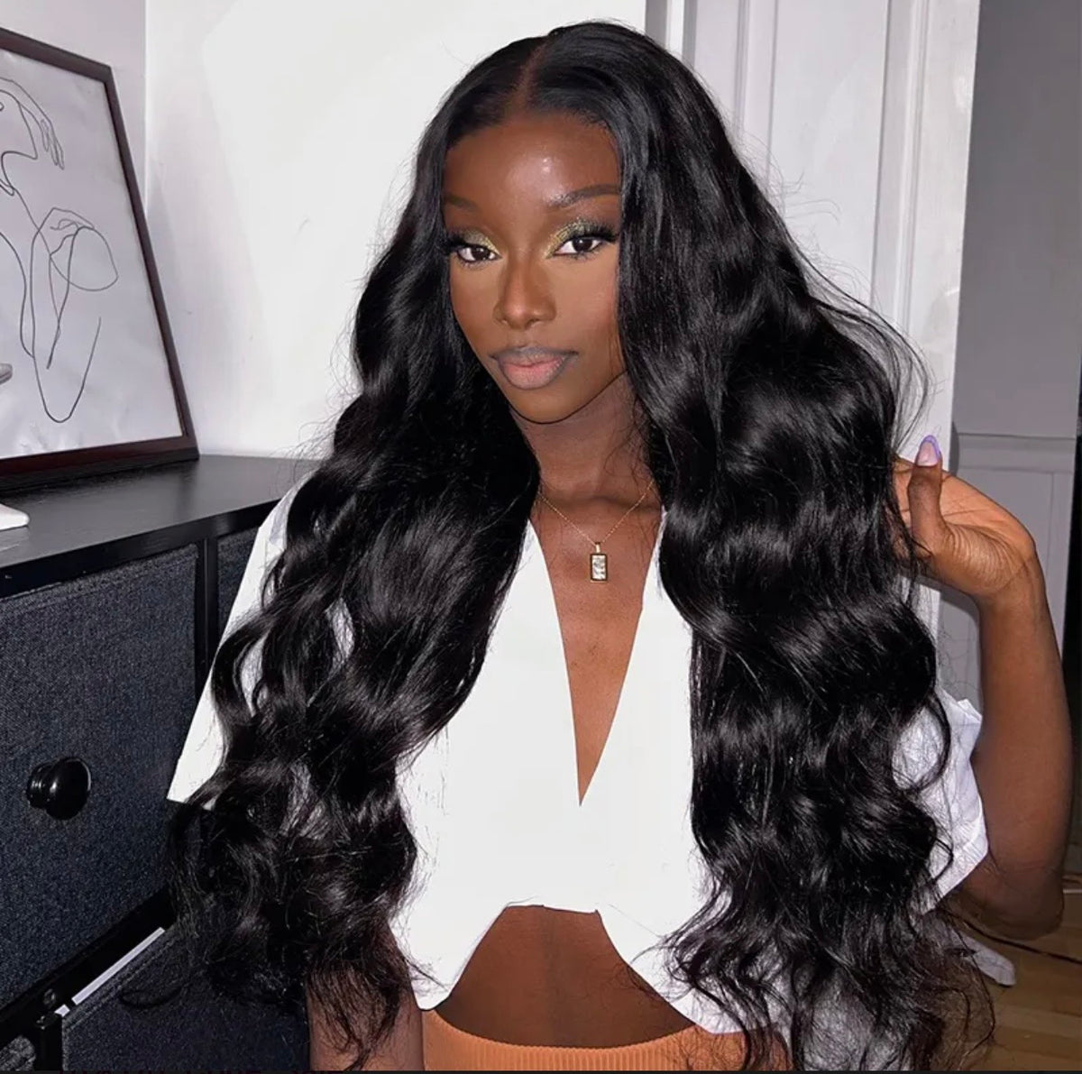 A woman with long, black, wavy hair wearing a white crop top, posing indoors with her hand touching her hair.