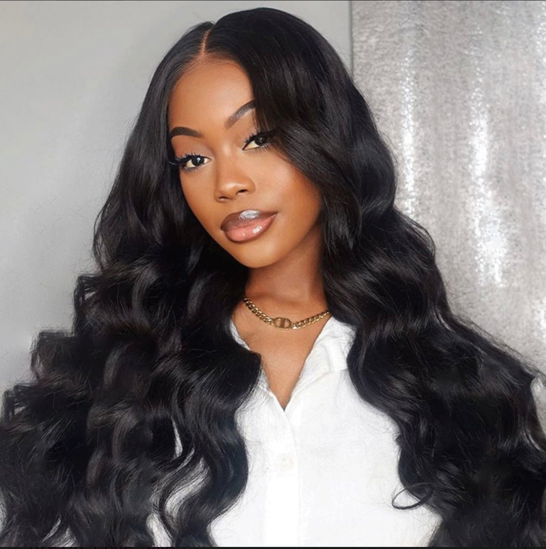 A woman with long, glossy, black wavy hair (likely a wig or hair extension) wearing a white collared shirt and a gold chain necklace, set against a gray and silver backdrop.