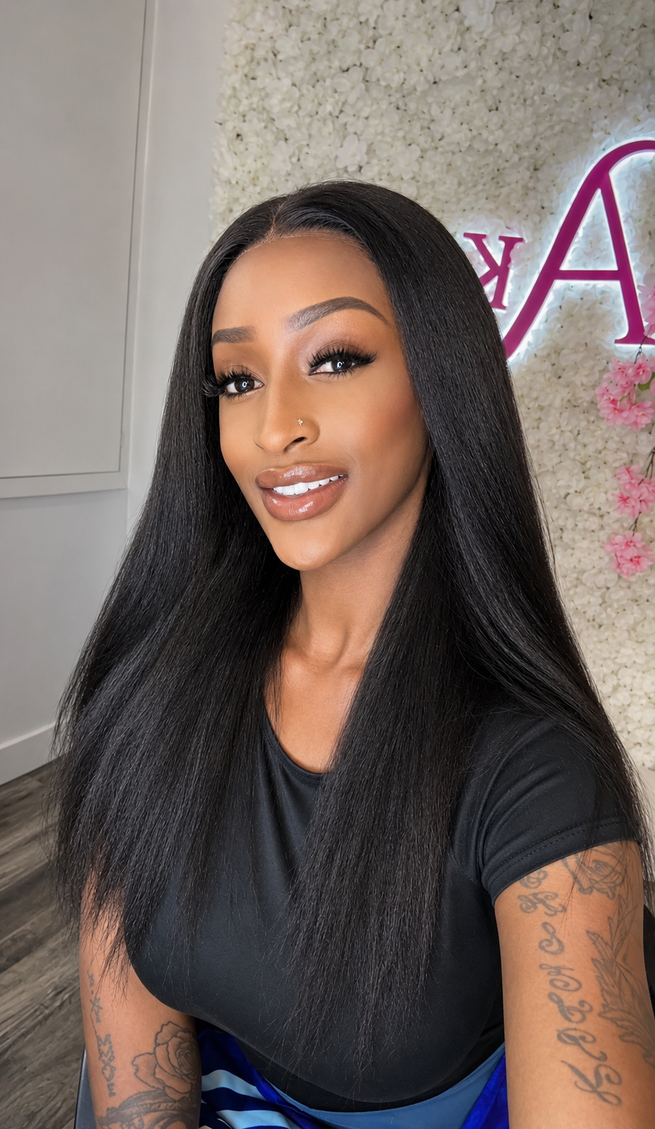 A woman with long, straight, black hair wearing a black t-shirt, posing indoors in front of a textured wall with a pink logo.