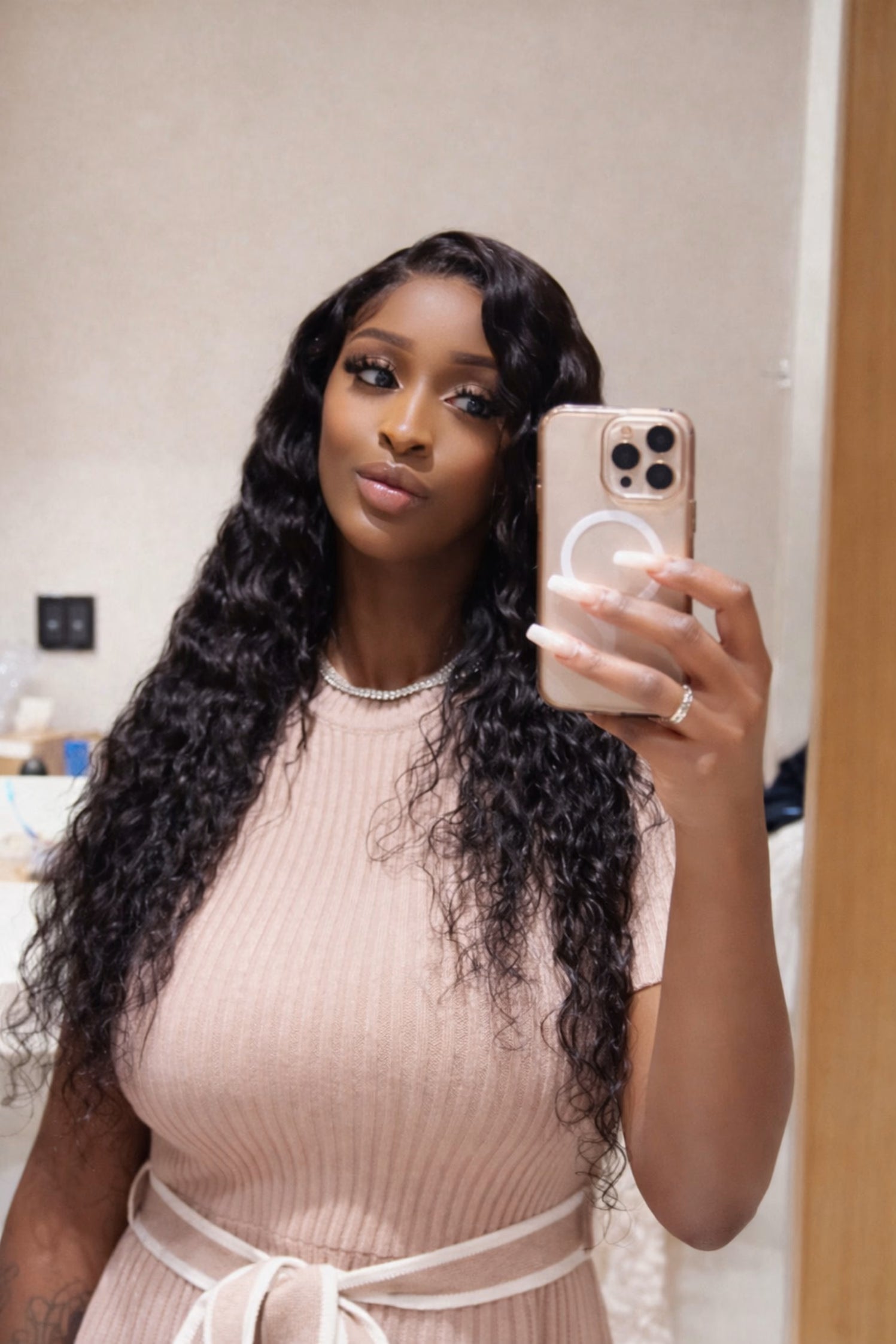 Woman taking a mirror selfie with long hair and a beige dress.