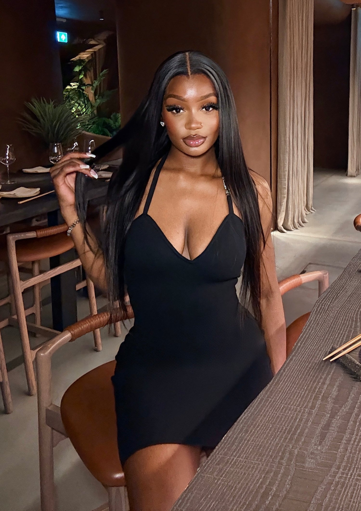 A woman with long, straight black hair wearing a black mini dress with a deep V-neckline, posing in a modern indoor setting with warm lighting.