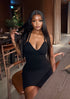 A woman with long, straight black hair wearing a black mini dress with a deep V-neckline, posing in a modern indoor setting with warm lighting.