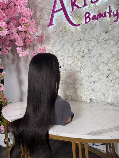 A long, straight, glossy black wig displayed on a mannequin head, shown from the back in a salon-like setting with a white floral wall, pink cherry blossoms, and a marble table.