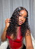A woman with long, dark, curly hair wearing a shiny red faux-leather top, posing in front of a gray curtain background.