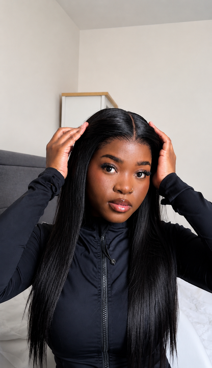 A woman with long, straight, black hair wearing a black zip-up jacket, posing indoors with her hands on her hair.